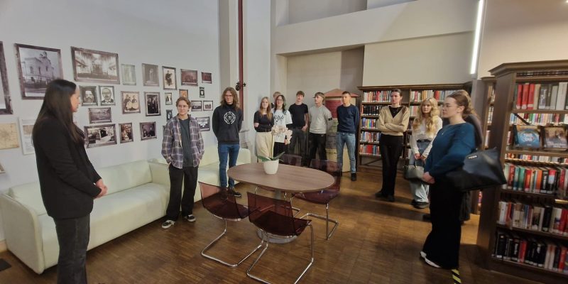 Jauniešu grupa stāv modernā bibliotēkas vai izstāžu zālē, klausoties sievietē, kas stāv priekšplānā pa kreisi. Sienas ir rotātas ar daudzām vecam fotogrāfijām, un labajā pusē ir augsti grāmatu plaukti.