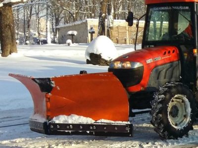 Sarkans traktors ar oranžu sniega lāpstu stāv uz sniegotas ceļa malas saulainā ziemas dienā.