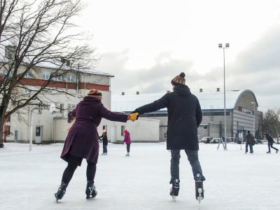 Pāris, turoties pie rokām, slido āra slidotavā ziemas dienā. Vīrietis valkā cepuri ar uzrakstu "LV", bet sieviete cepuri ar uzrakstu "SILVE". Fonā redzamas ēkas un citi slidotāji. Kuldīgas slidotava.