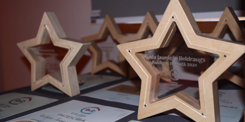 Vairāki koka zvaigznes formas apbalvojumi ar caurspīdīgiem centriem un sertifikāti uz galda. Uz priekšējā apbalvojuma ir uzraksts: "Gada jauniešu lieldraugs Kuldīgas novadā 2024".