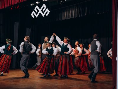 Folkloras dejotāji tradicionālos latviešu tērpos uzstājas uz skatuves. Sievietes valkā garus svītrainus svārkus, zaļas vestes un vainagus, vīrieši – pelēkas bikses un vestes. Aiz dejotājiem pie melnās sienas redzama liela balta Jumja zīme.