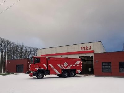 Sarkana ugunsdzēsēju mašīna ar baltām svītrām, uzrakstu "BLAŅES", emblēmu un numuru "402" novietota pie moderna ugunsdzēsēju depo ēkas, uz kuras fasādes redzami lieli sarkani cipari "112". Zeme ir apsnigusi.