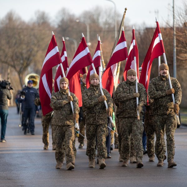 Vairāki vīrieši militārās maskēšanās formās soļo pa ielu, nesot paceltus Latvijas karogus. Fonā redzams orķestris