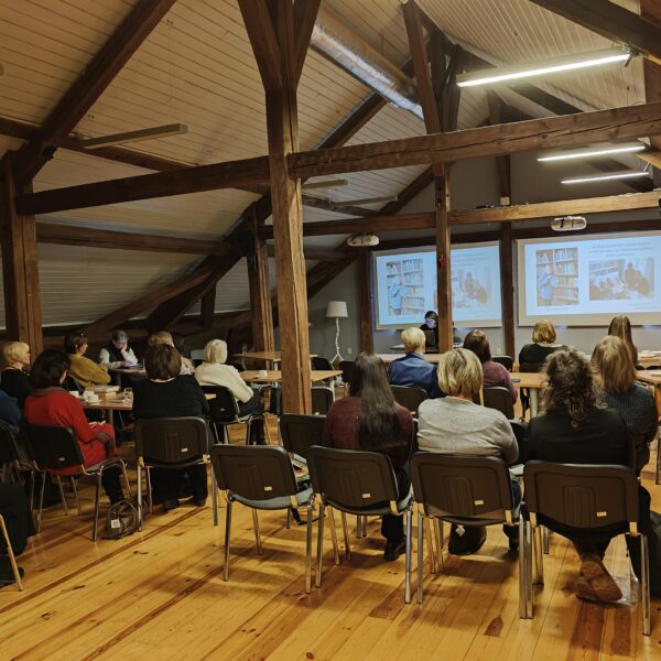 Bēniņu telpa ar atsegtām koka sijām, kurā pieaugušo grupa sēž krēslos, vērojot prezentāciju. Priekšā redzami divi projektoru ekrāni, kuros attēloti grāmatu plaukti un cilvēki, kas lasa vai strādā pie galdiem.