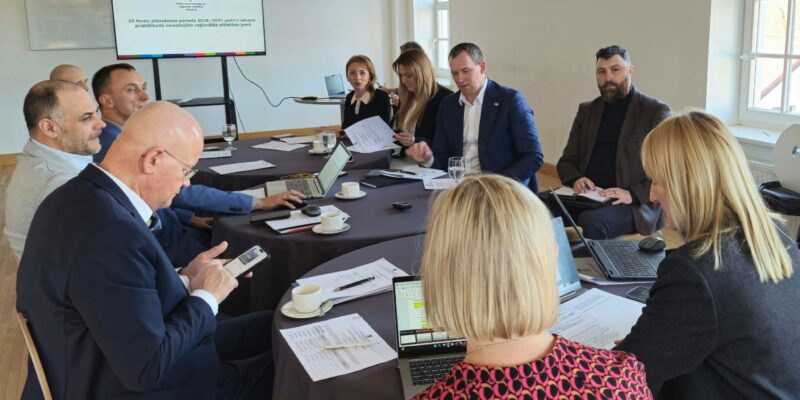 Sanāksme ar aptuveni desmit cilvēkiem, kas sēž pie liela galda, daži strādā ar klēpjdatoriem un pārskata dokumentus. Liels ekrāns rāda tekstu "ES fondu plānošanas periods 2028-2034 gadam Latvijā: priekšlikumi investīcijām reģionālās attīstības jomā".