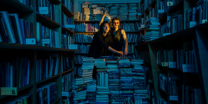 Divi smaidīgi vīrieši, viens ar tumšām saulesbrillēm un tetovētām rokām, blāvi apgaismotā bibliotēkā veido mūziku pie dīdžeja aprīkojuma "Pioneer DJ", kas novietots uz augstas grāmatu kaudzes. Viņu apkārtnē ir plaukti pilni ar grāmatām un grīda ir nokrauta ar papildu grāmatu kaudzēm.